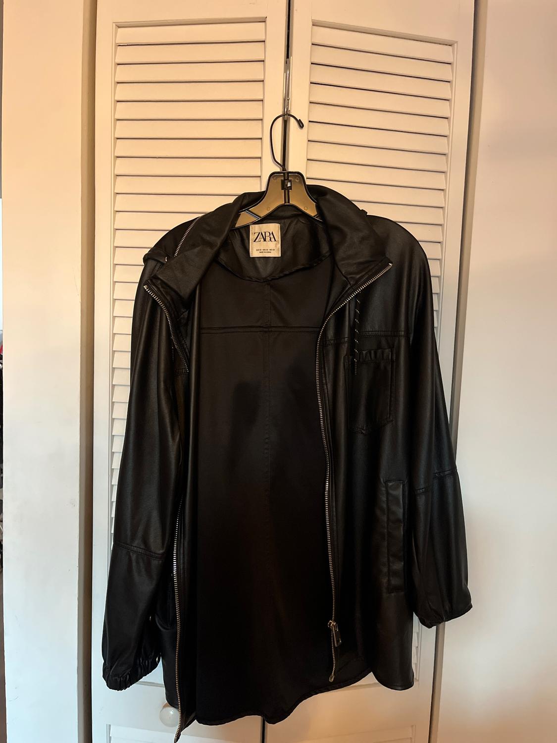 Black jacket hanging on a hanger in front of a white door with blinds. Visible label with the text "ZARA".