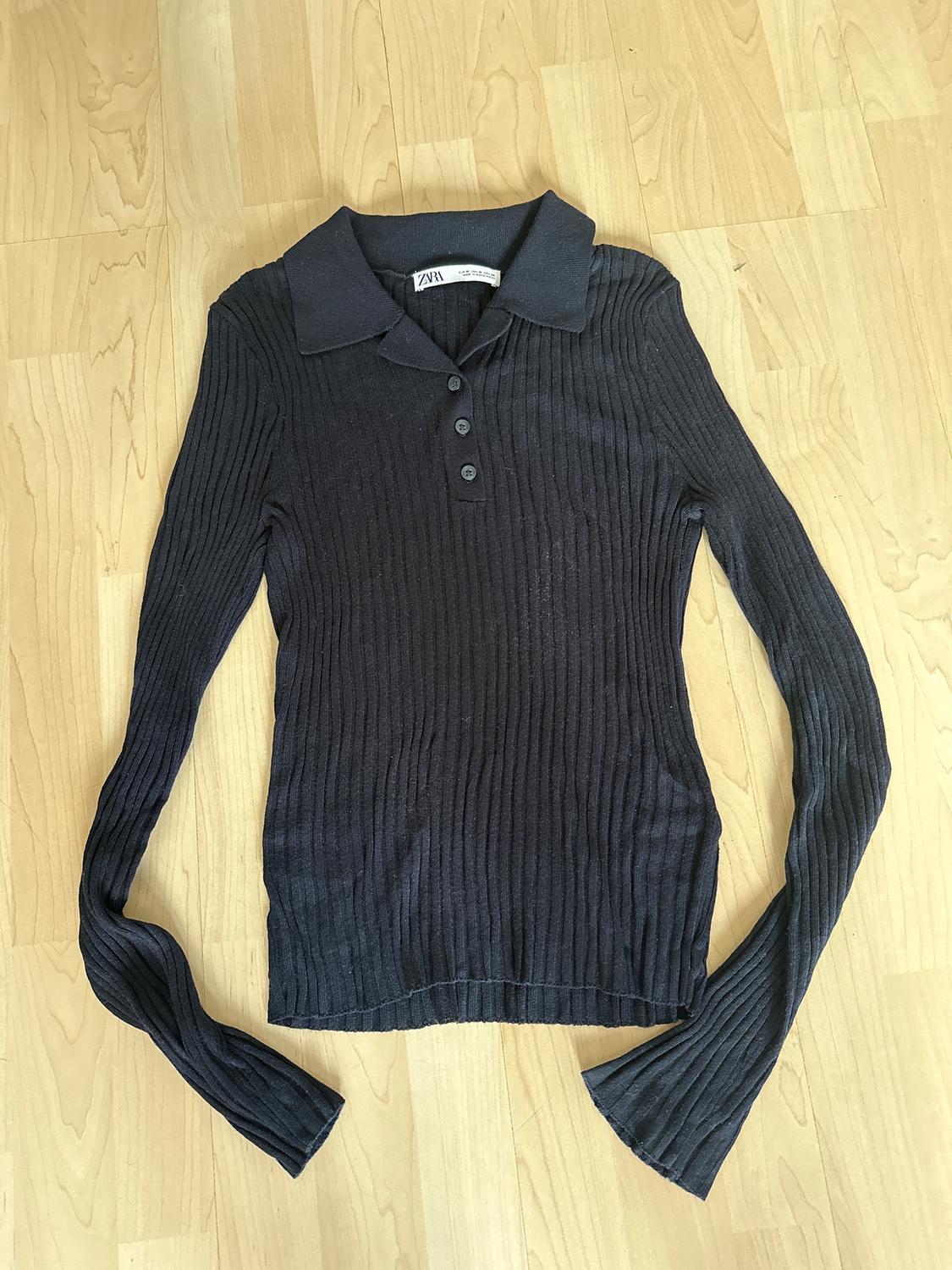 Black long-sleeved ribbed jumper with a collar and buttons, on a light wooden floor. Visible label: "Zara".
