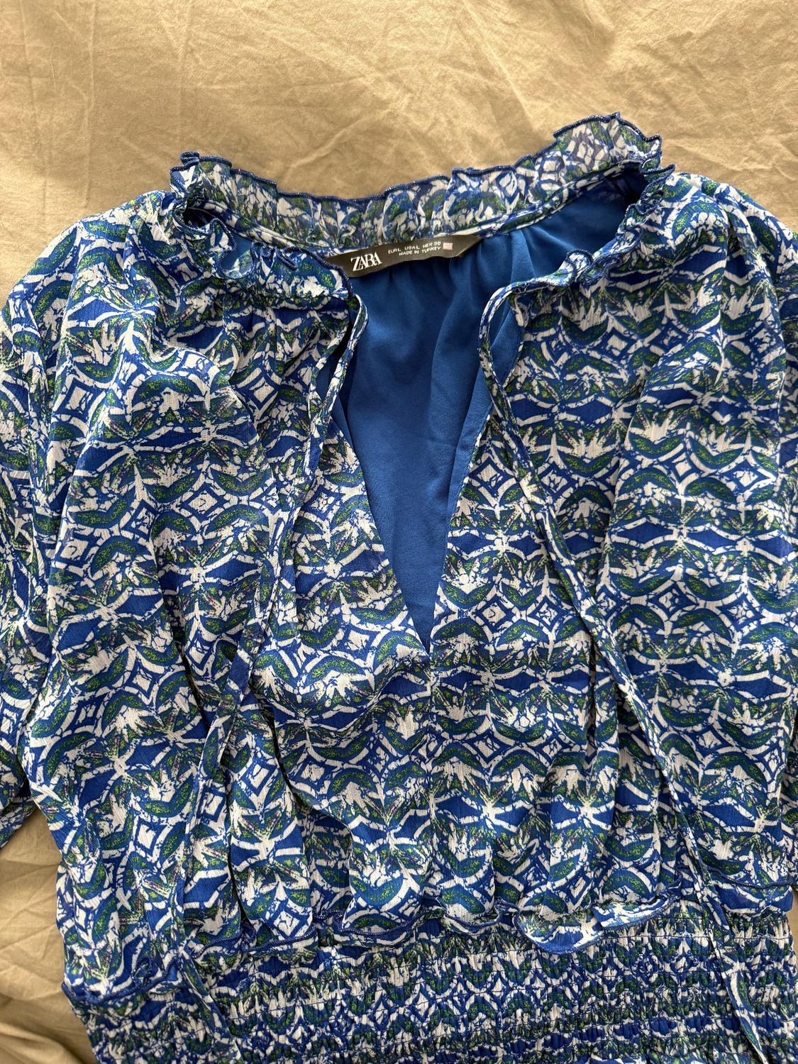 Blue dress with a geometric print in white and green, with a ruffled collar and visible ZARA tag. Beige background.