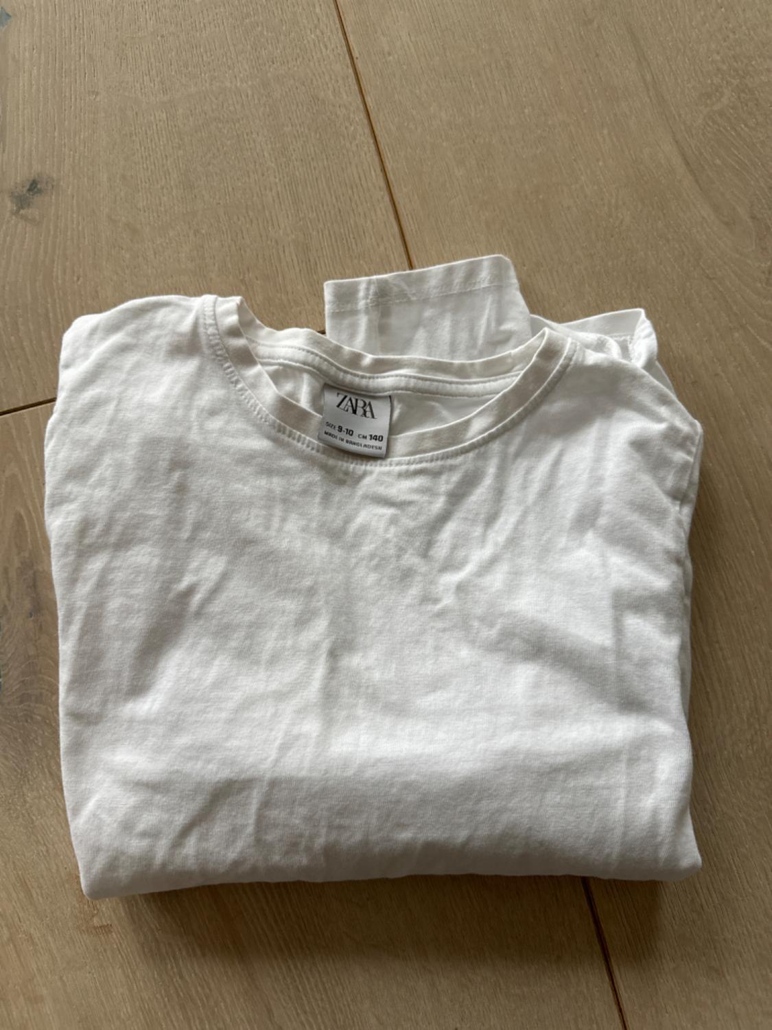 White T-shirt folded on a light wooden surface. The label reads "ZARA" and size "9-10 years, 140 cm".