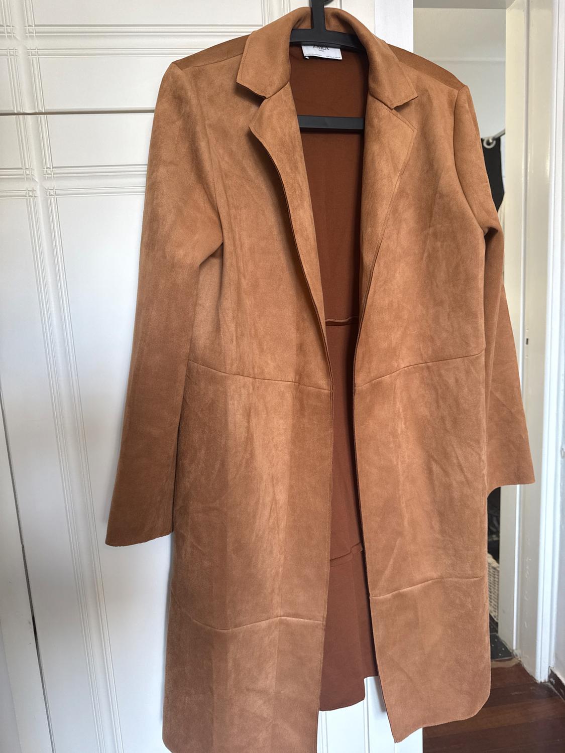 Light brown coat hanging on a black hanger, over a white door. Soft fabric and simple design with lapels.