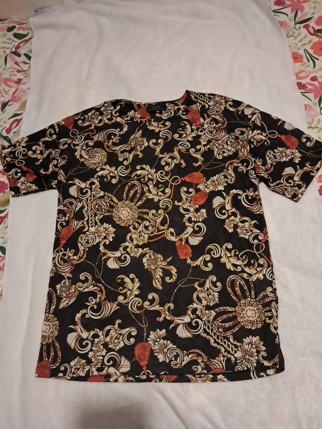 Black short-sleeved T-shirt with a gold baroque print and red tassels, label with ZARA text, laid out on white fabric.