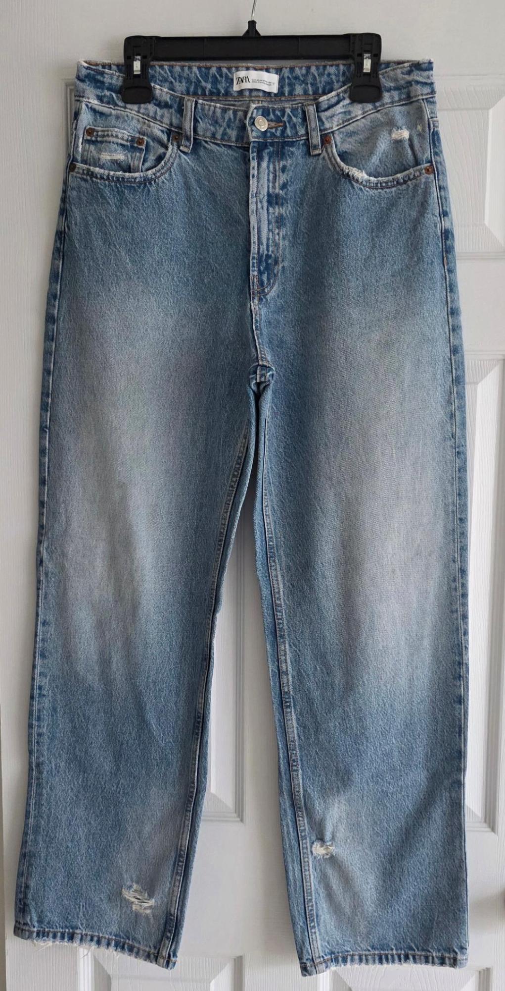 Worn blue jeans hanging on a black hanger in front of a white door. Visible tag: "ZARA".
