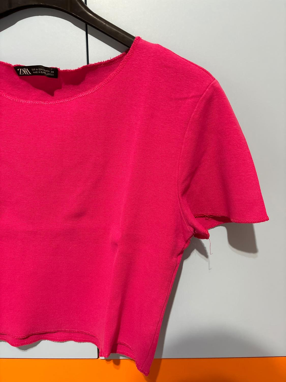 Φωτεινό ροζ T-shirt κρεμασμένο σε μαύρη κρεμάστρα, πάνω σε λευκό και πορτοκαλί φόντο. Ορατή ετικέτα στο λαιμό.