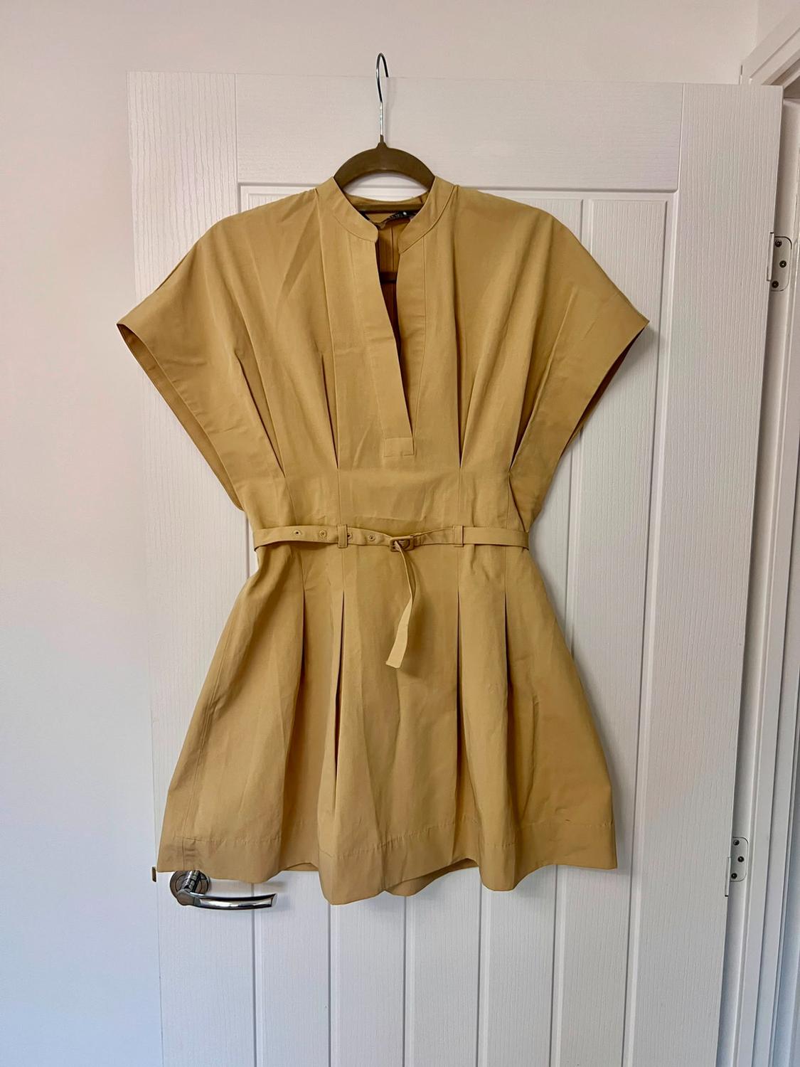 Mustard-coloured short dress with a V-neck, short sleeves, pleats and a thin belt, hanging on a hanger on a white door.