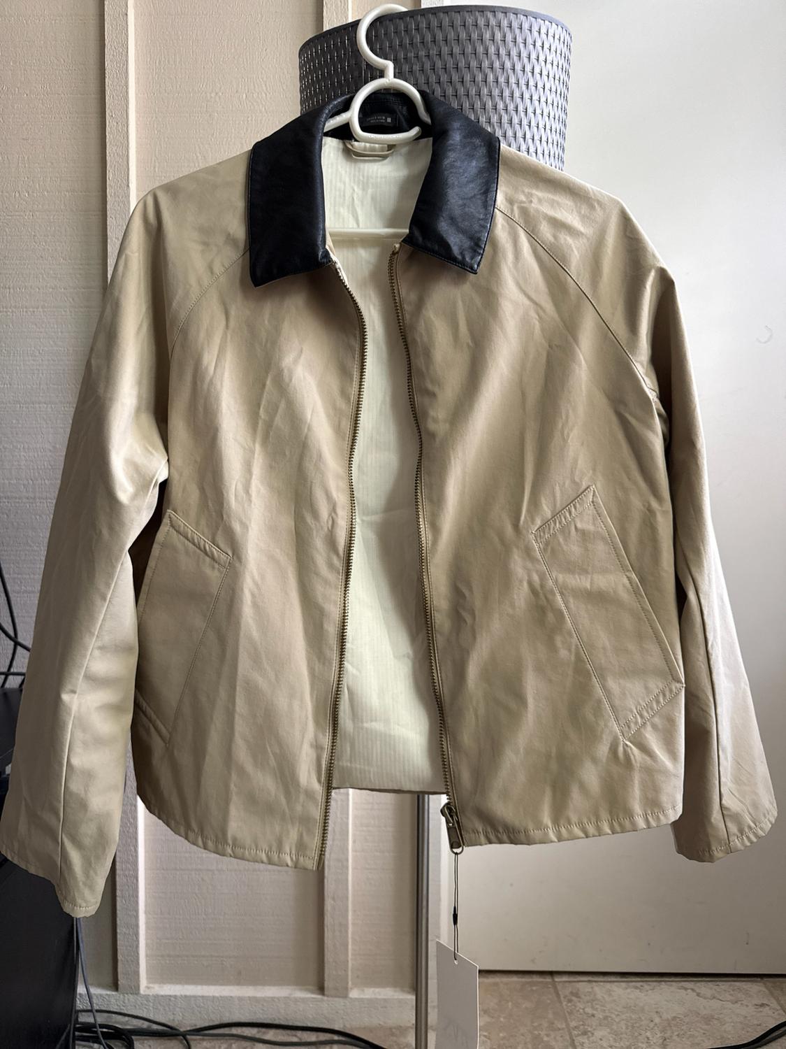 Beige jacket with a contrasting black collar and open front zipper, hanging on a white hanger in an indoor space.