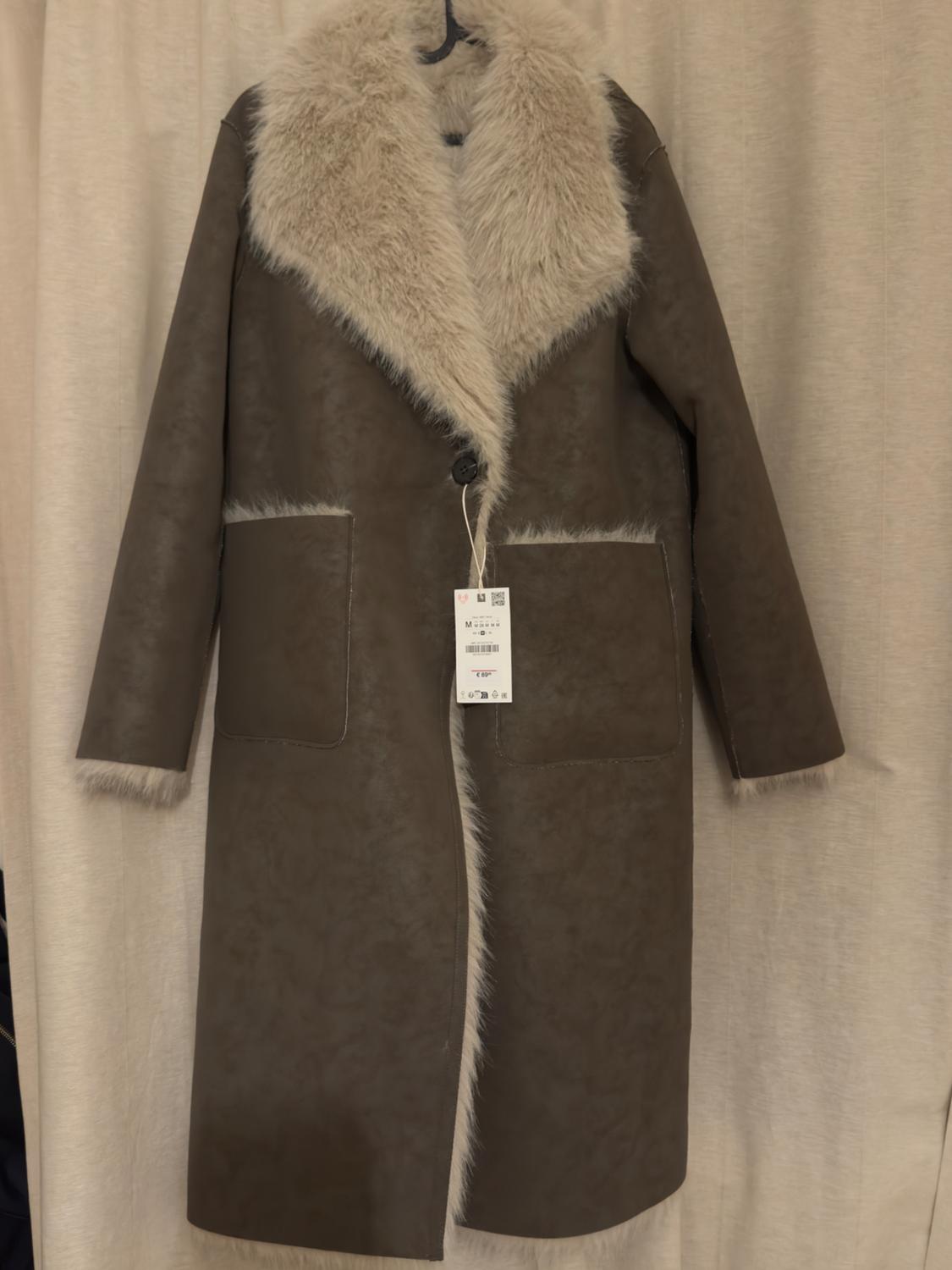 Long brown coat with a beige fur collar and trim, hanging on a hanger. Label visible on the front. Beige curtain background.