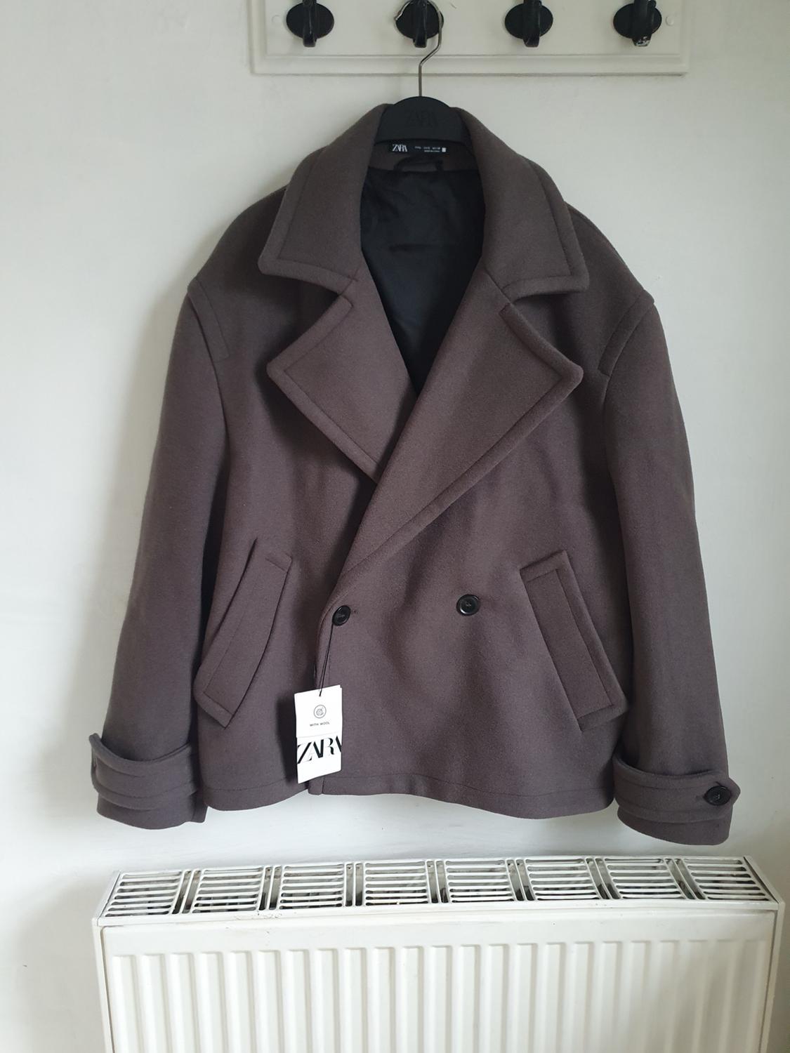 Dark grey coat hanging on a black hanger over a white radiator. Visible label with the text "ZARA".