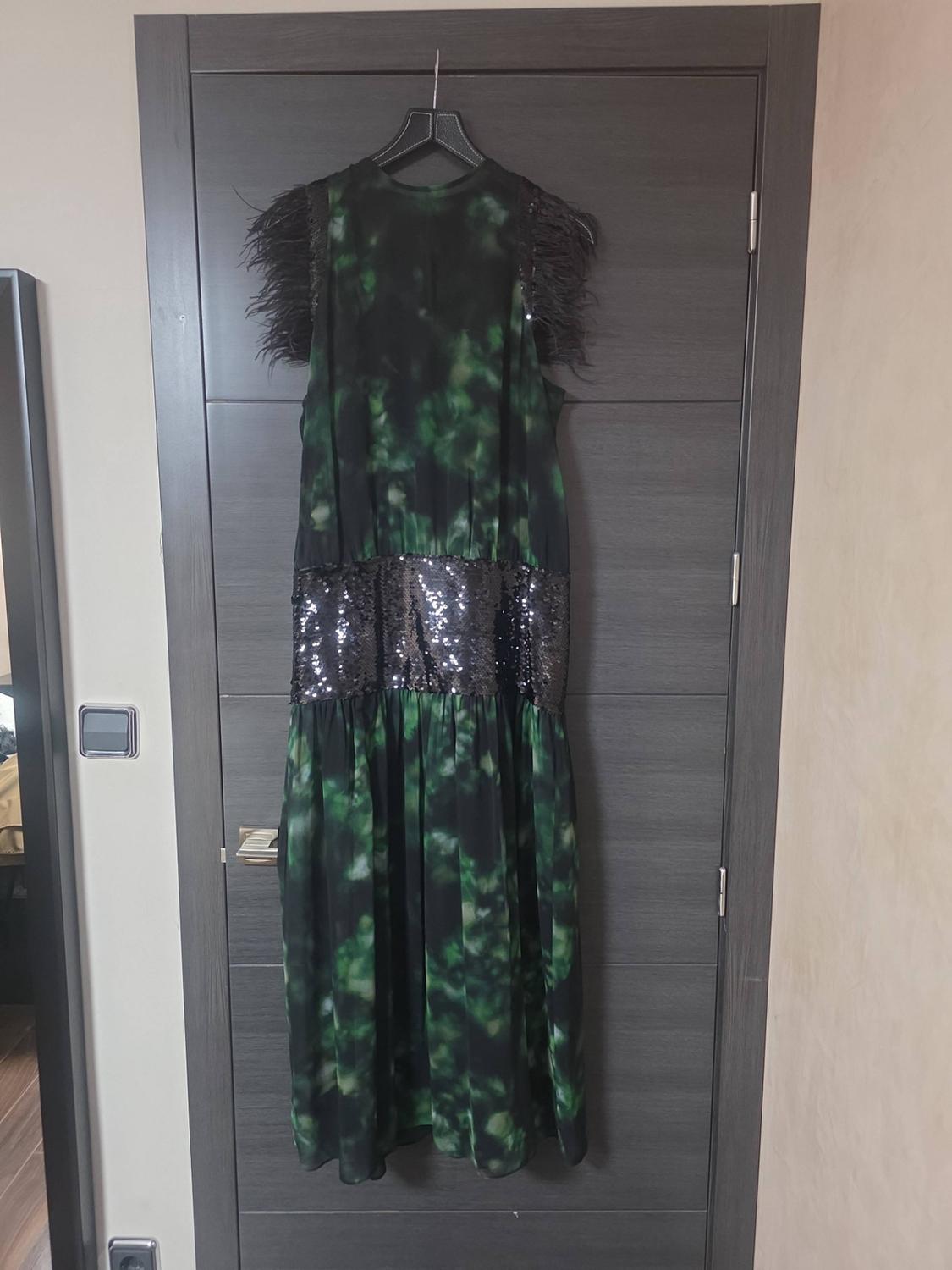 Green and black patterned maxi dress, with feathers on the shoulders and a black sequined waist, hanging on a wooden door.