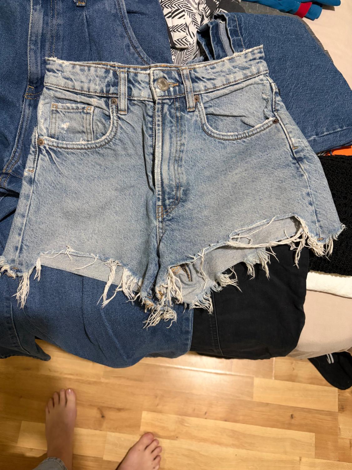 Distressed denim shorts on a bed with other denim garments. Wooden floor and bare feet visible.