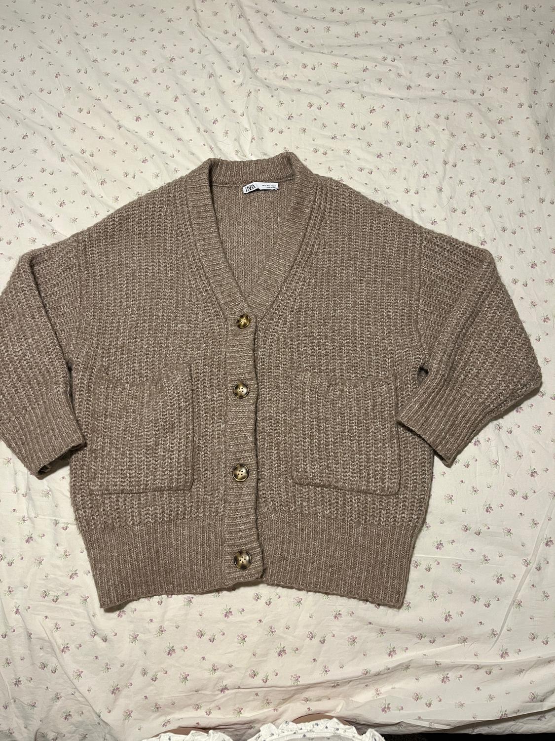 Brown knit cardigan with gold buttons on a white floral-print quilt. Visible tag on the collar.