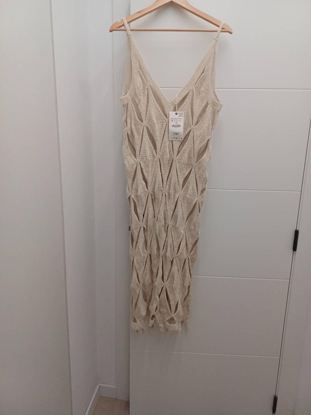 Beige knitted maxi dress with an openwork diamond pattern and thin straps, hanging on a wooden hanger against a white door.