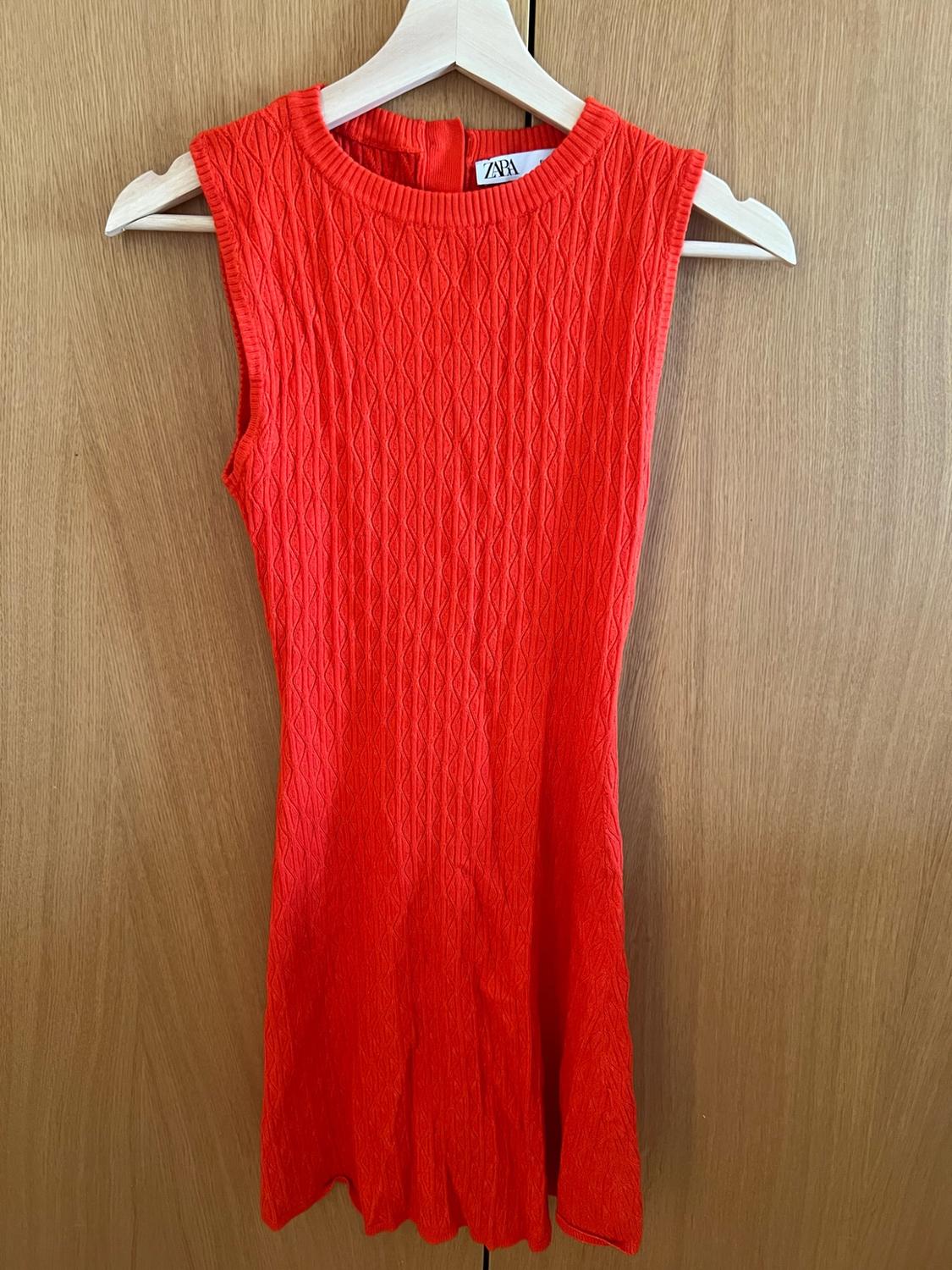 Sleeveless red knitted dress with diamond texture, on a white hanger against a wooden background. Label with the text ZARA.