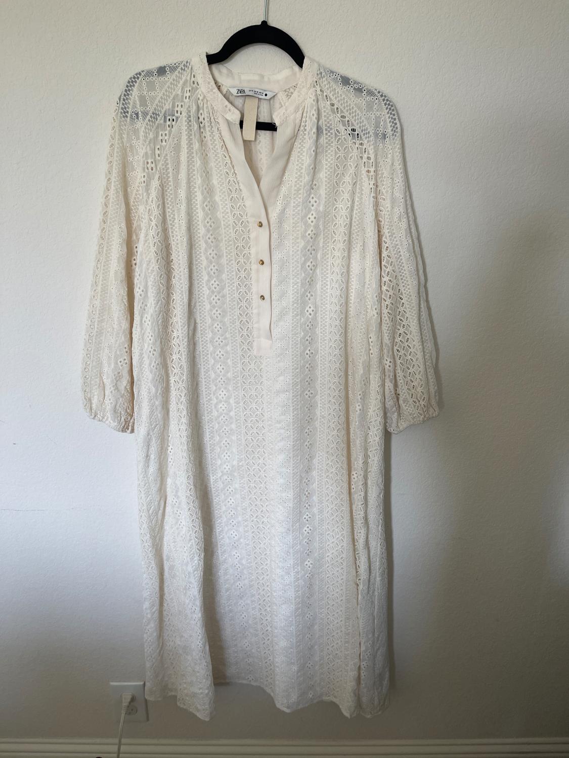 White lace dress with three-quarter sleeves and buttons on the chest, hanging on a black clothes hanger against a white wall.