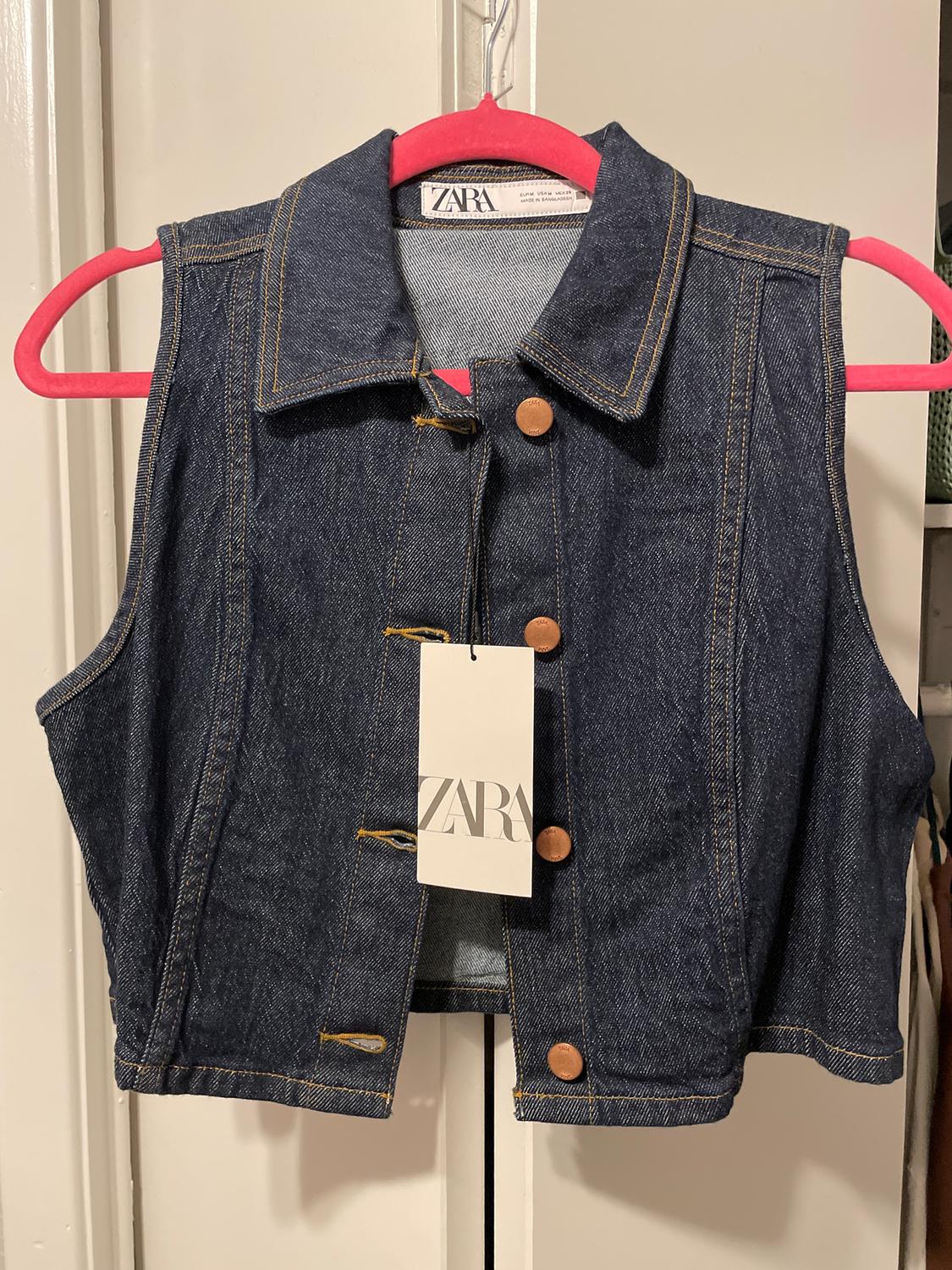 Blue sleeveless denim waistcoat with brown buttons, hanging on a red hanger. Visible label with the text "ZARA".