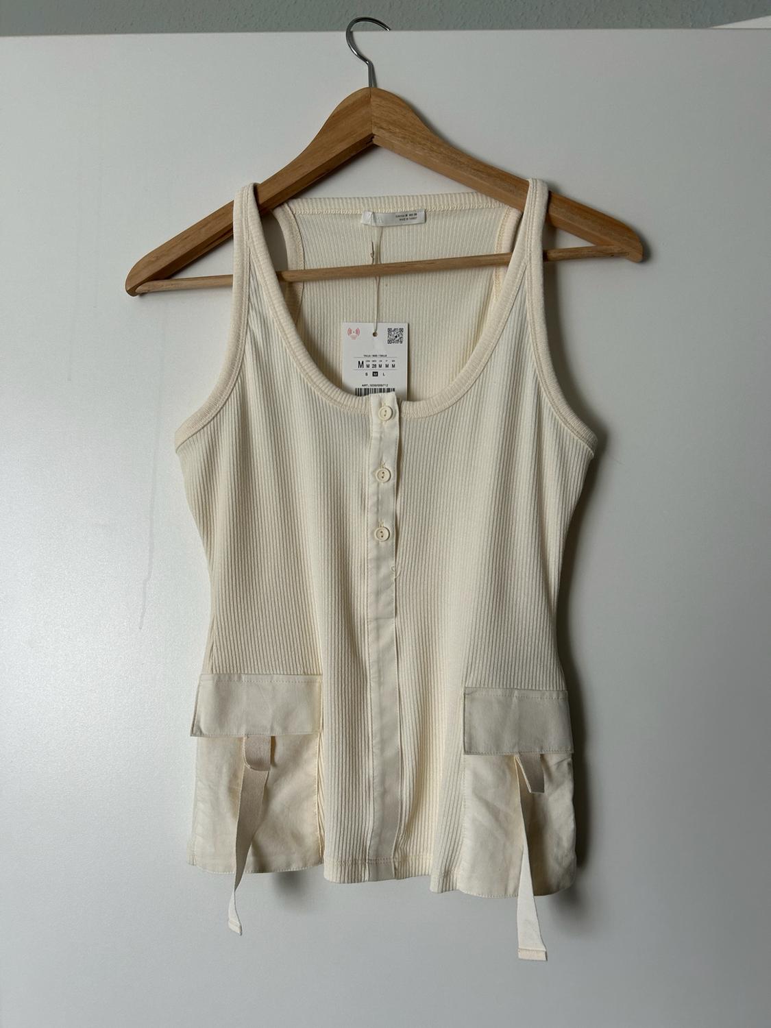 Cream ribbed vest top with buttons and front patch pockets with straps, hanging on a wooden hanger against a white background.