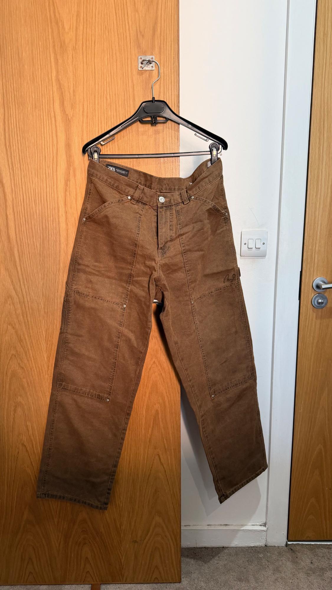 Brown trousers with front panels hanging on a wooden door. Label with the text ZARA. Casual setting with a white wall and a light switch.