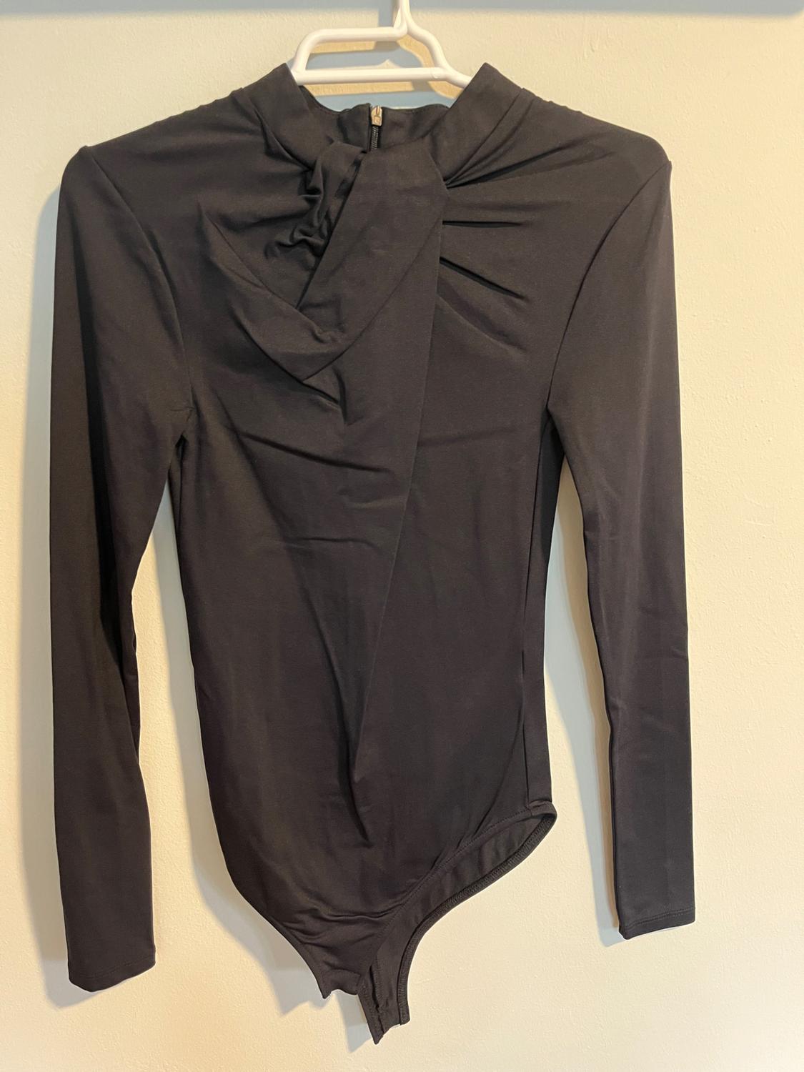 Black long-sleeved high-neck bodysuit with pleat detail on the chest, hanging on a white hanger against a light background.