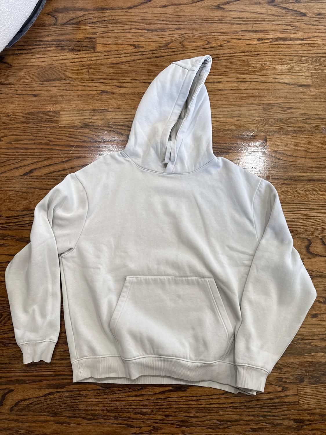 Light gray hooded sweatshirt, long sleeves, and a front pocket, spread out on a wooden floor.