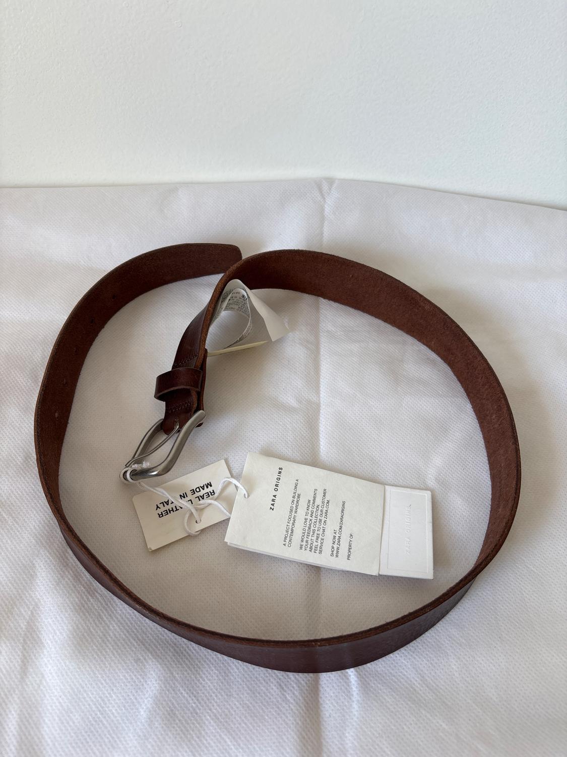 Brown leather belt with metal buckle on a white background. Visible labels: "REAL LEATHER MADE IN ITALY" and "ZARA ORIGINS".