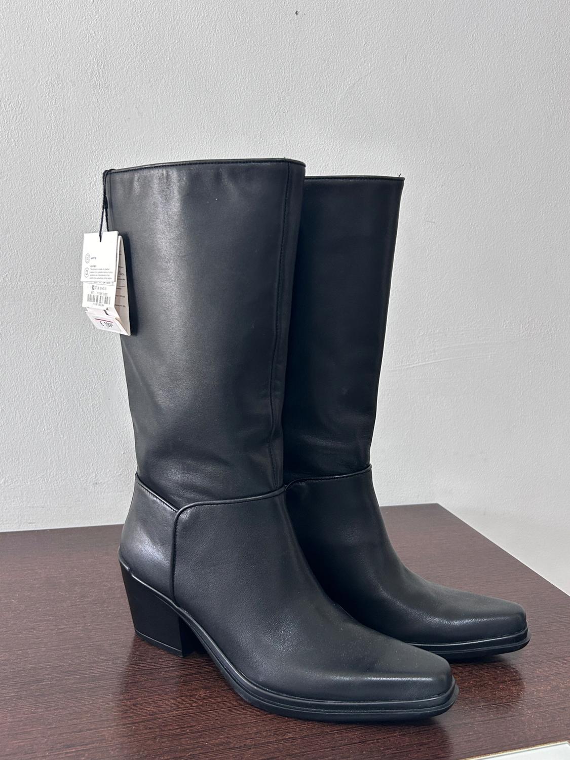 Black mid-heel boots on a dark wooden surface, with a visible hang tag on one of them.