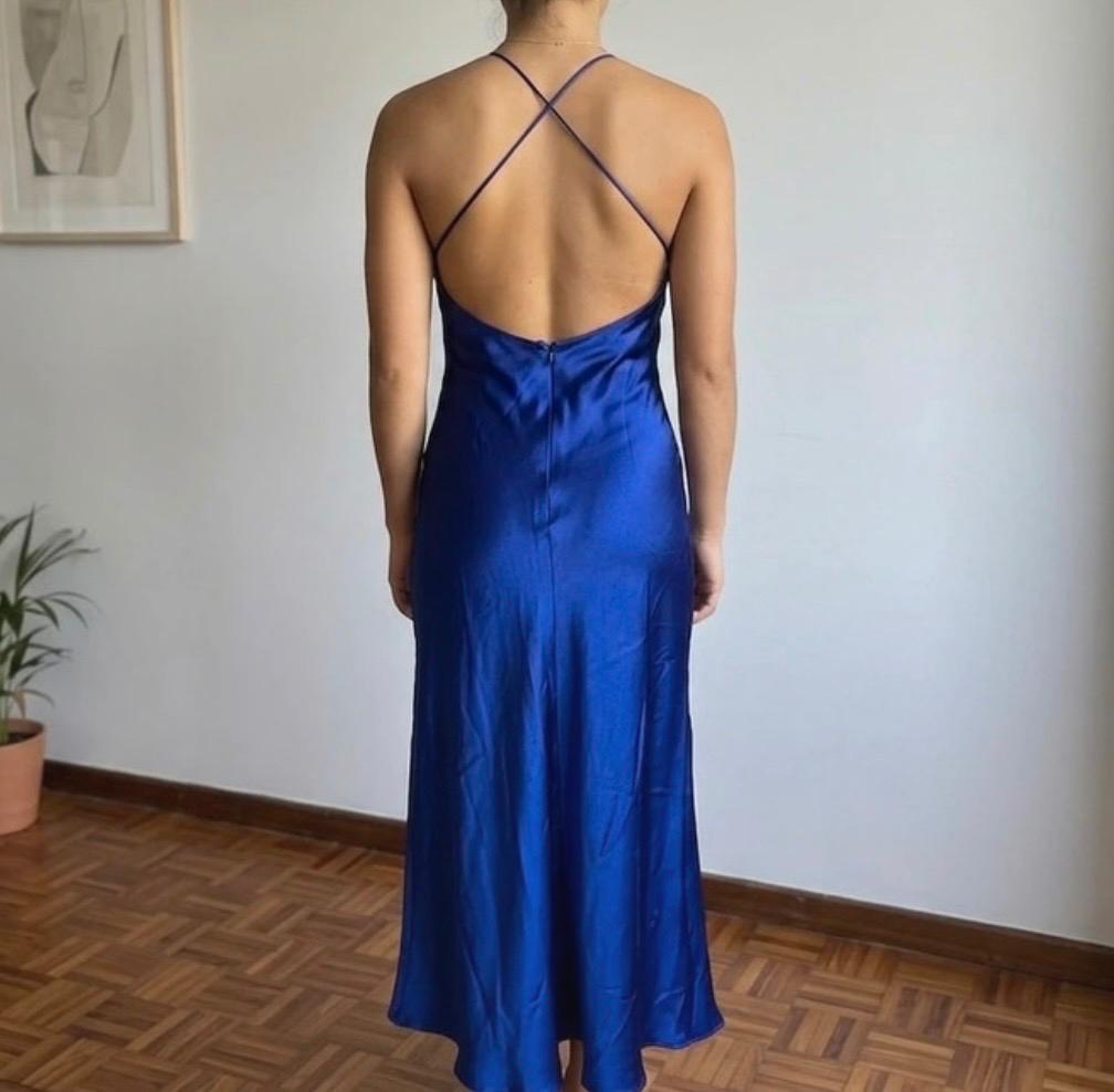 Model from the back wearing a long satin blue dress with thin crossed straps and an open back, indoors with a wooden floor.