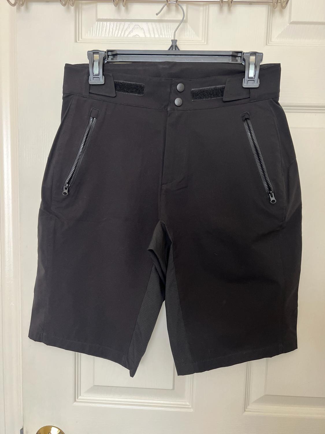 Black shorts hanging on a hanger over a white door. Its front zip pockets and two-button fastening stand out.