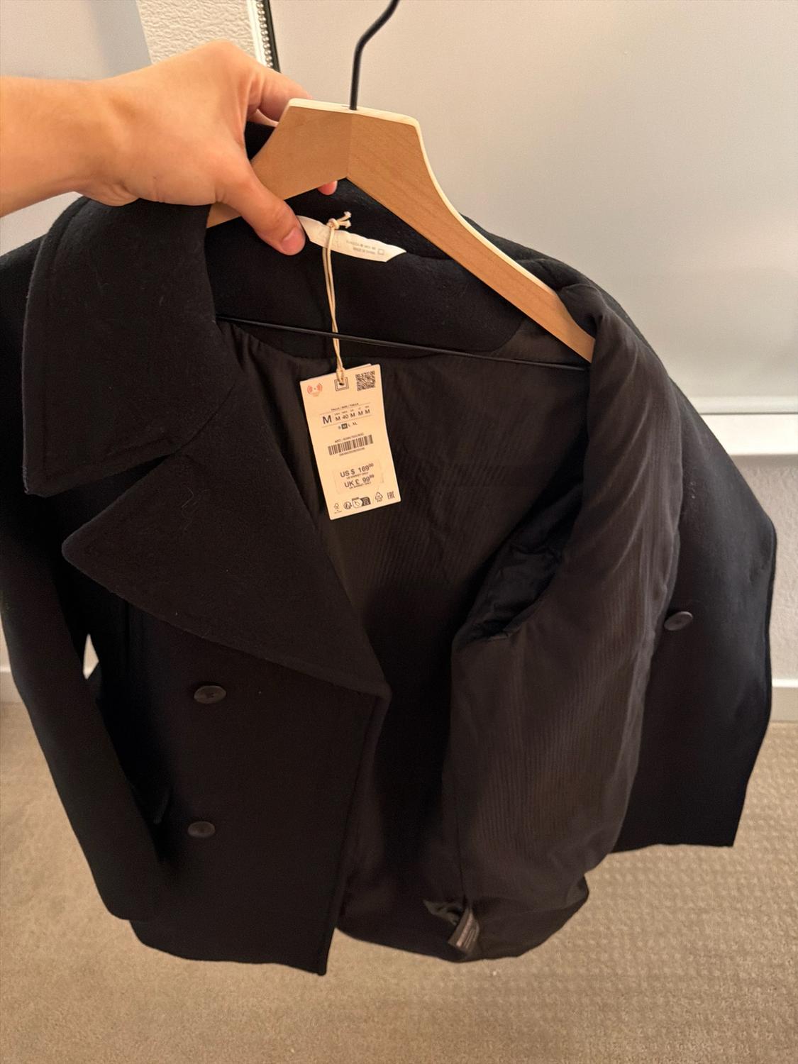 Hand holding a hanger with an open black double-breasted coat. Tag with text: M M 40 M M M US $ 169.00 UK £ 99.99.