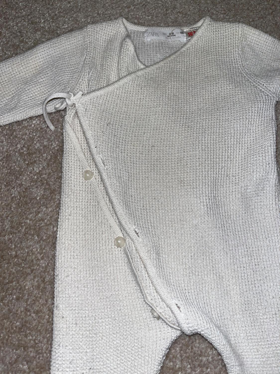 Knitted cream baby all-in-one with buttons and a side bow, on a brown rug. Visible label on the collar.