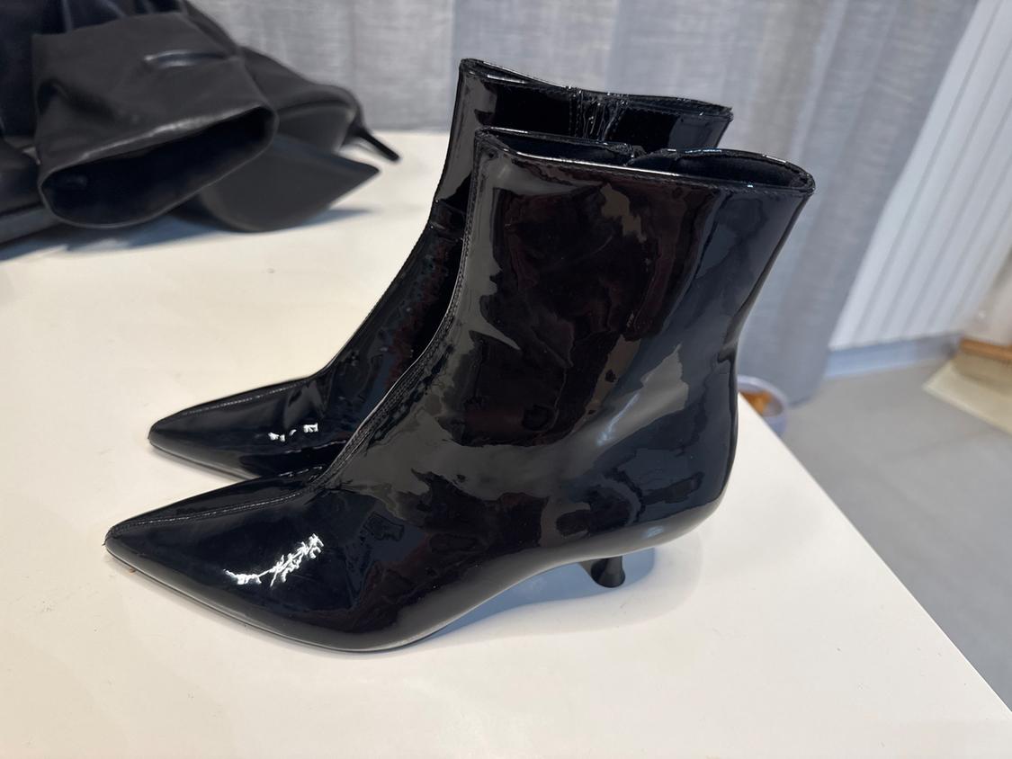 Black patent leather ankle boots with a low heel and pointed toe on a white table, with a grey curtain background.