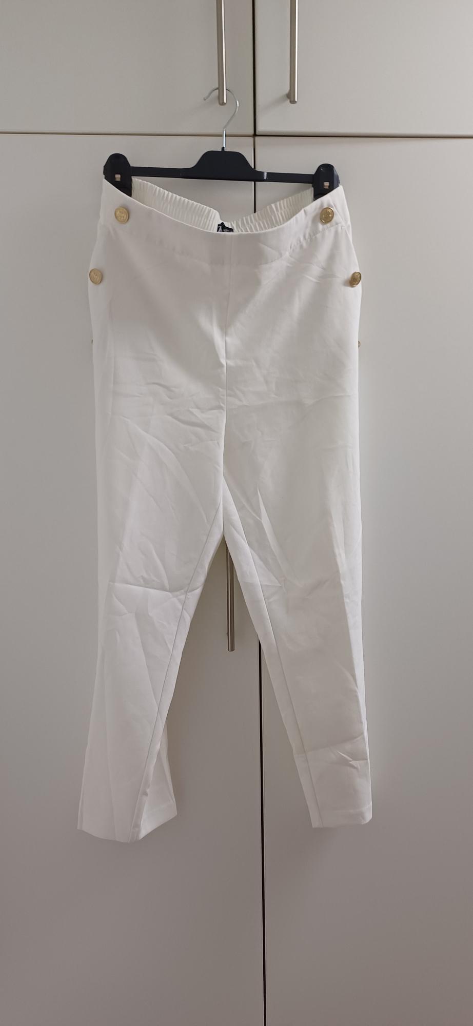 White trousers hanging on a black hanger, with gold buttons on the sides, in front of a white wardrobe.