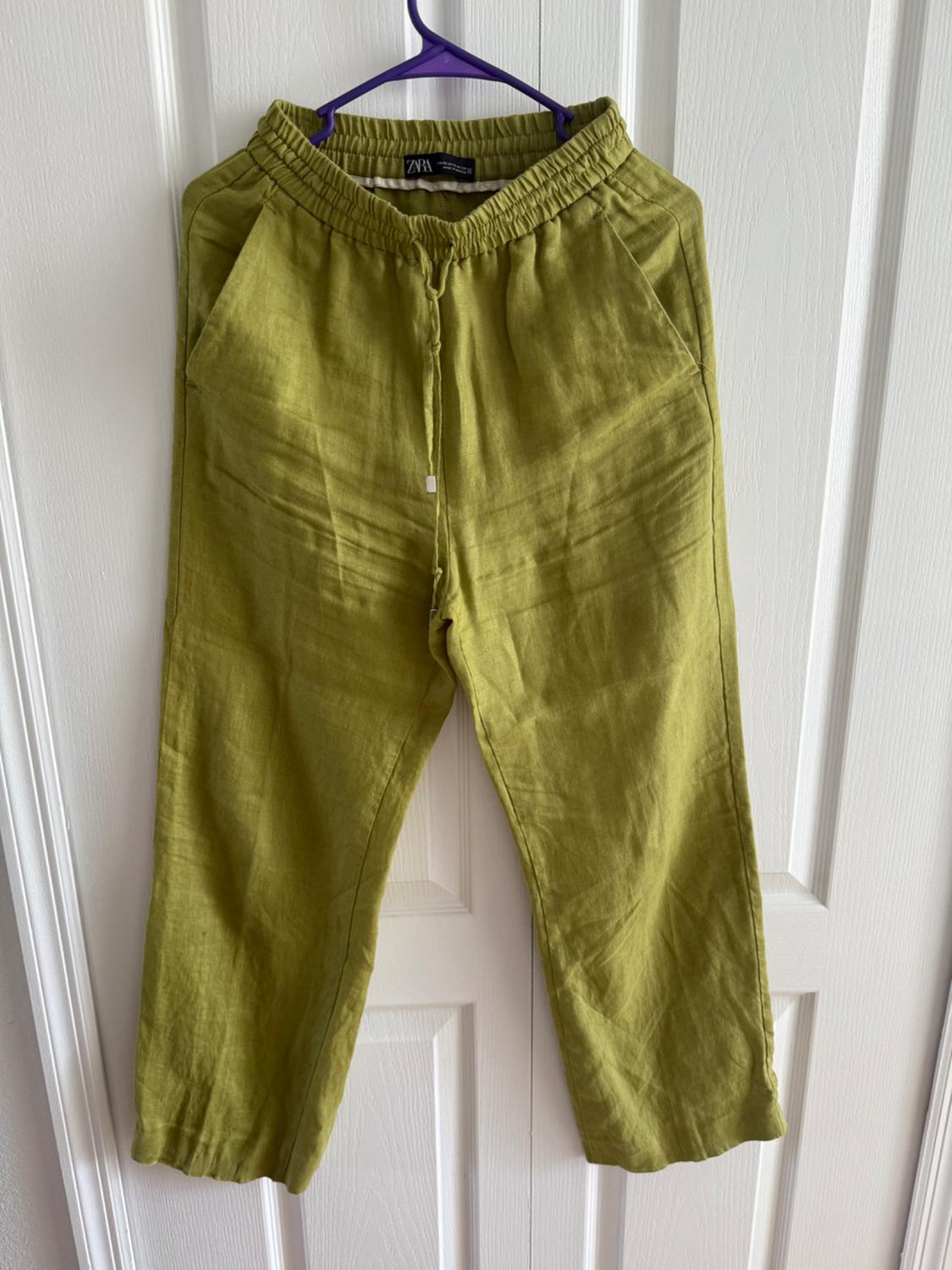 Green pants hanging on a purple hanger in front of a white door. They have an elastic waistband and side pockets.