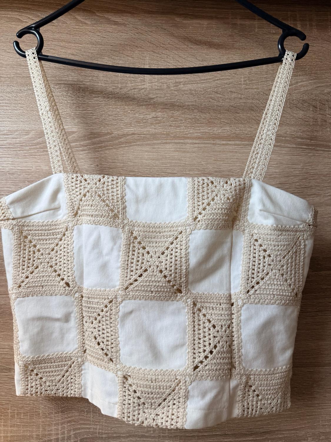 White strappy top with a beige fabric and crochet checked design, hanging on a black hanger against a wooden background.