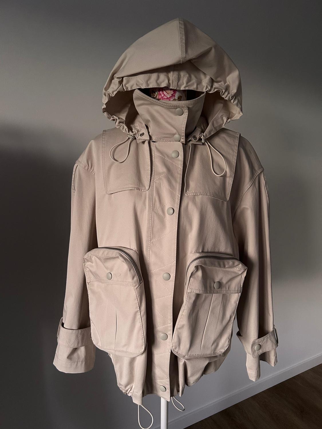 Beige jacket with a hood and large front pockets, displayed on a mannequin against a dark grey background.