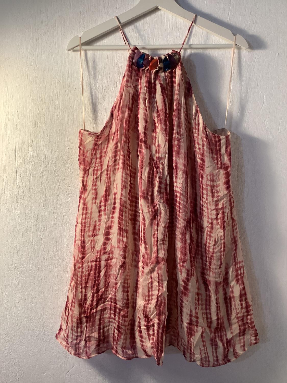 Αμάνικο φόρεμα με στάμπα tie-dye σε κόκκινους και λευκούς τόνους, κρεμασμένο σε μια λευκή κρεμάστρα πάνω από έναν ανοιχτόχρωμο τοίχο.