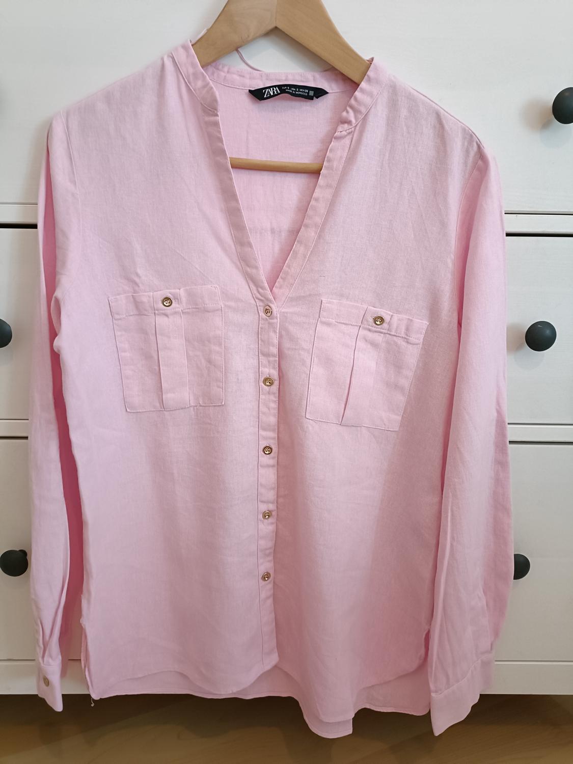 Pink long-sleeved button-down shirt with chest pockets, hanging on a wooden hanger in front of a white chest of drawers. ZARA label.