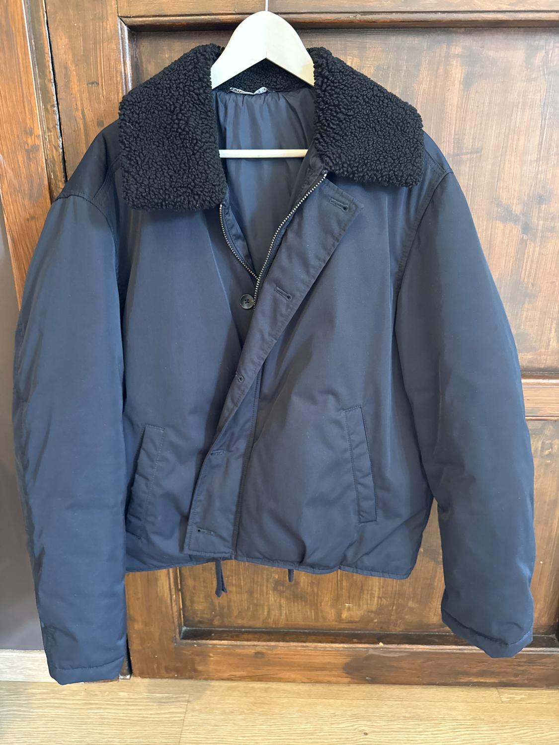 Dark blue jacket with a black shearling collar, hanging on a white hanger in front of a wooden door.