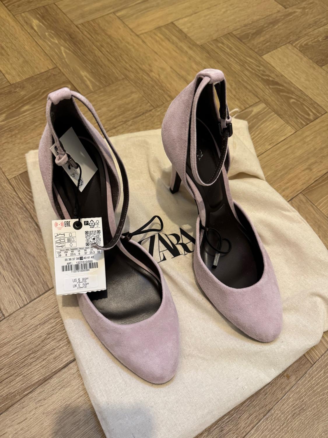 Lilac suede high-heeled shoes on a bag with ZARA text on a wooden floor. Label with text US $ 89.90 UK £ 59.99.