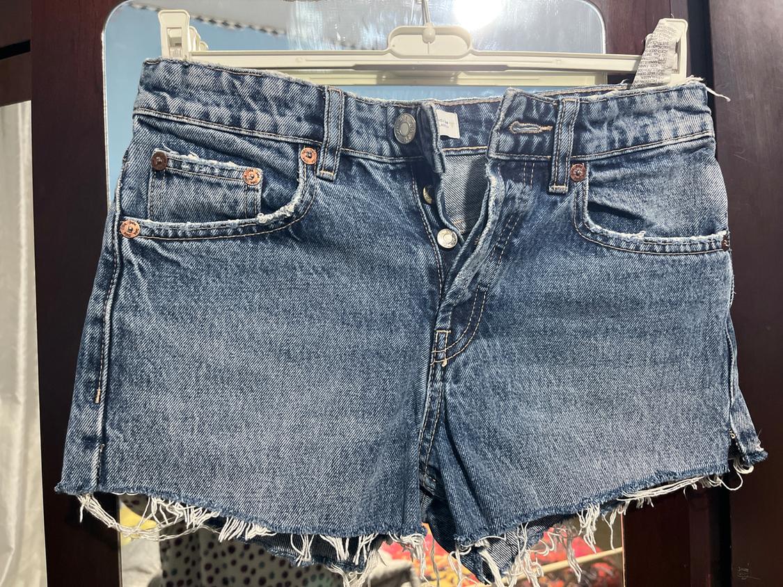 Blue denim shorts with frayed hem, hanging on a white hanger against a dark wooden background.