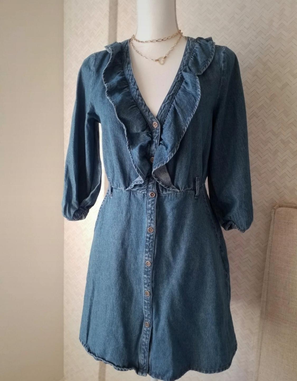 Mannequin wearing a blue denim dress with ruffles and front buttons. Light background with subtle pattern. Gold necklace around the neck.