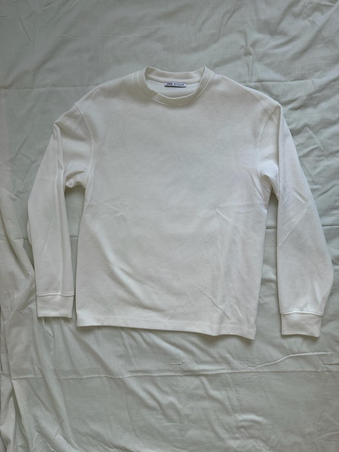 White long-sleeved T-shirt with ZARA label, laid out on a crumpled white sheet. Relaxed style flat lay.