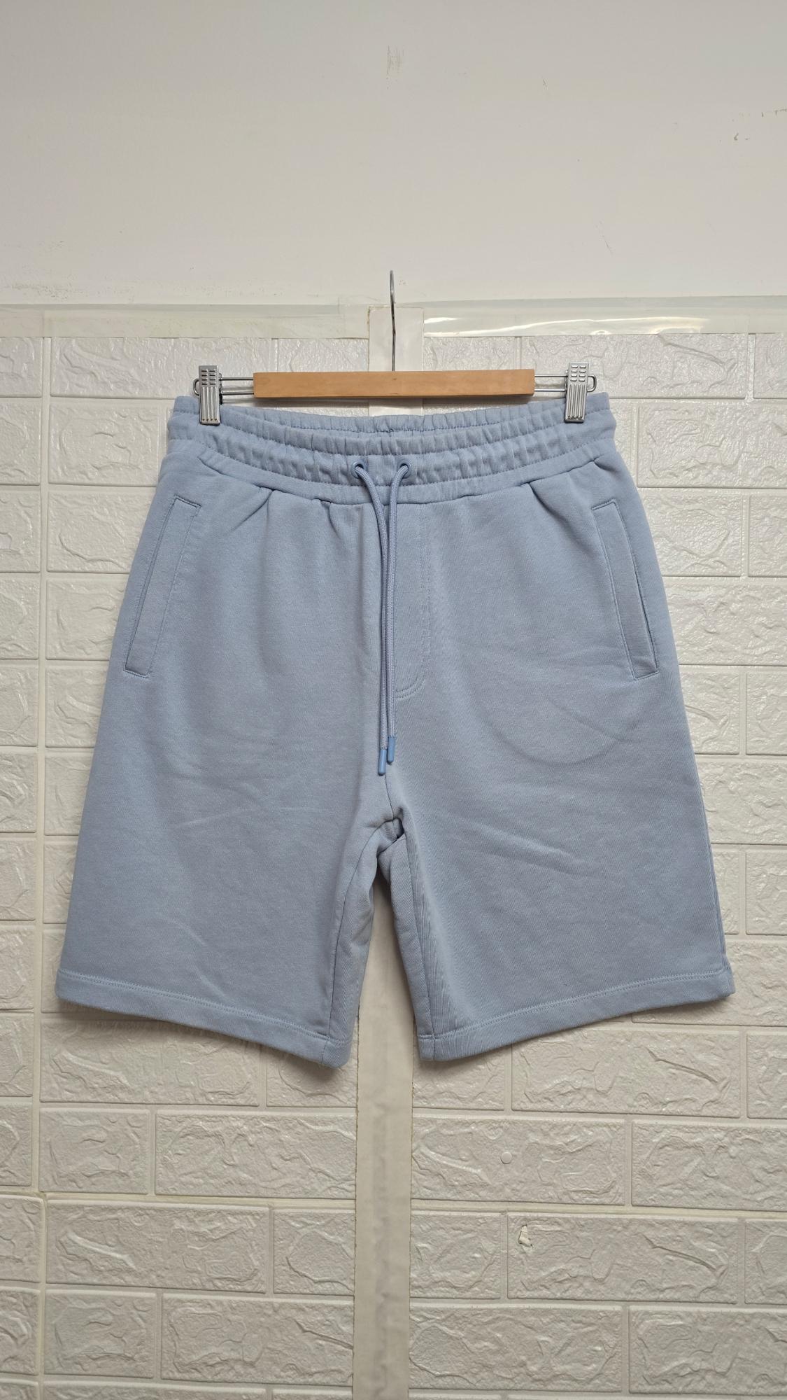 Light blue shorts hanging on a wooden hanger, with adjustable drawstring and side pockets, against a white wall background.