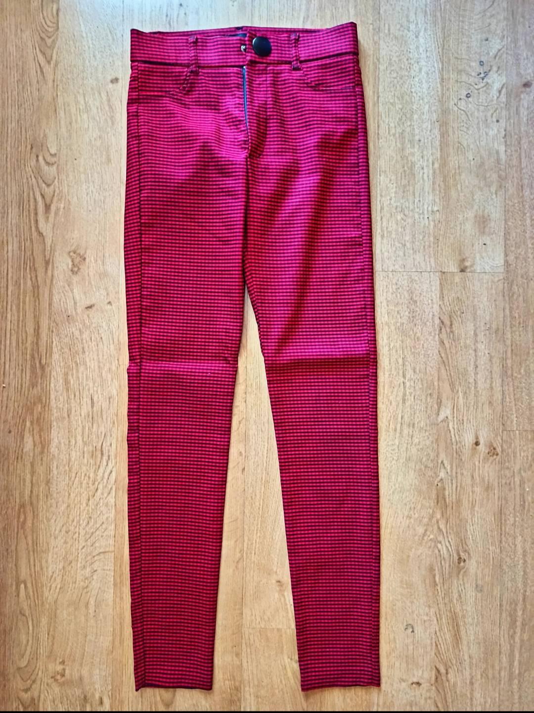 Slim-fit trousers with a small red and black check pattern, featuring a dark button at the waist, laid out on a light wooden floor.