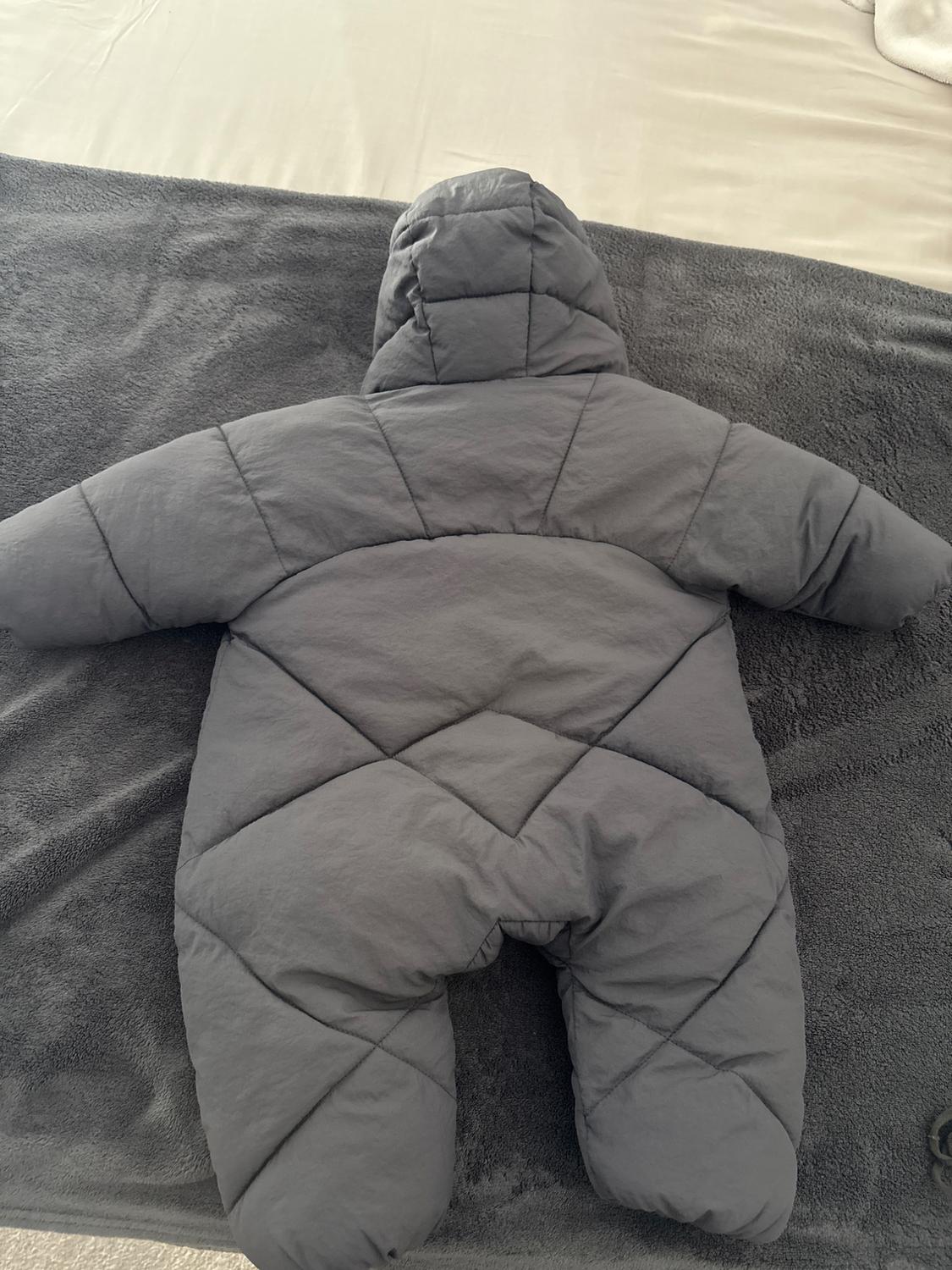 Grey padded jumpsuit with hood, diamond-quilted stitching design, laid on a grey blanket on a bed with light-coloured sheets.