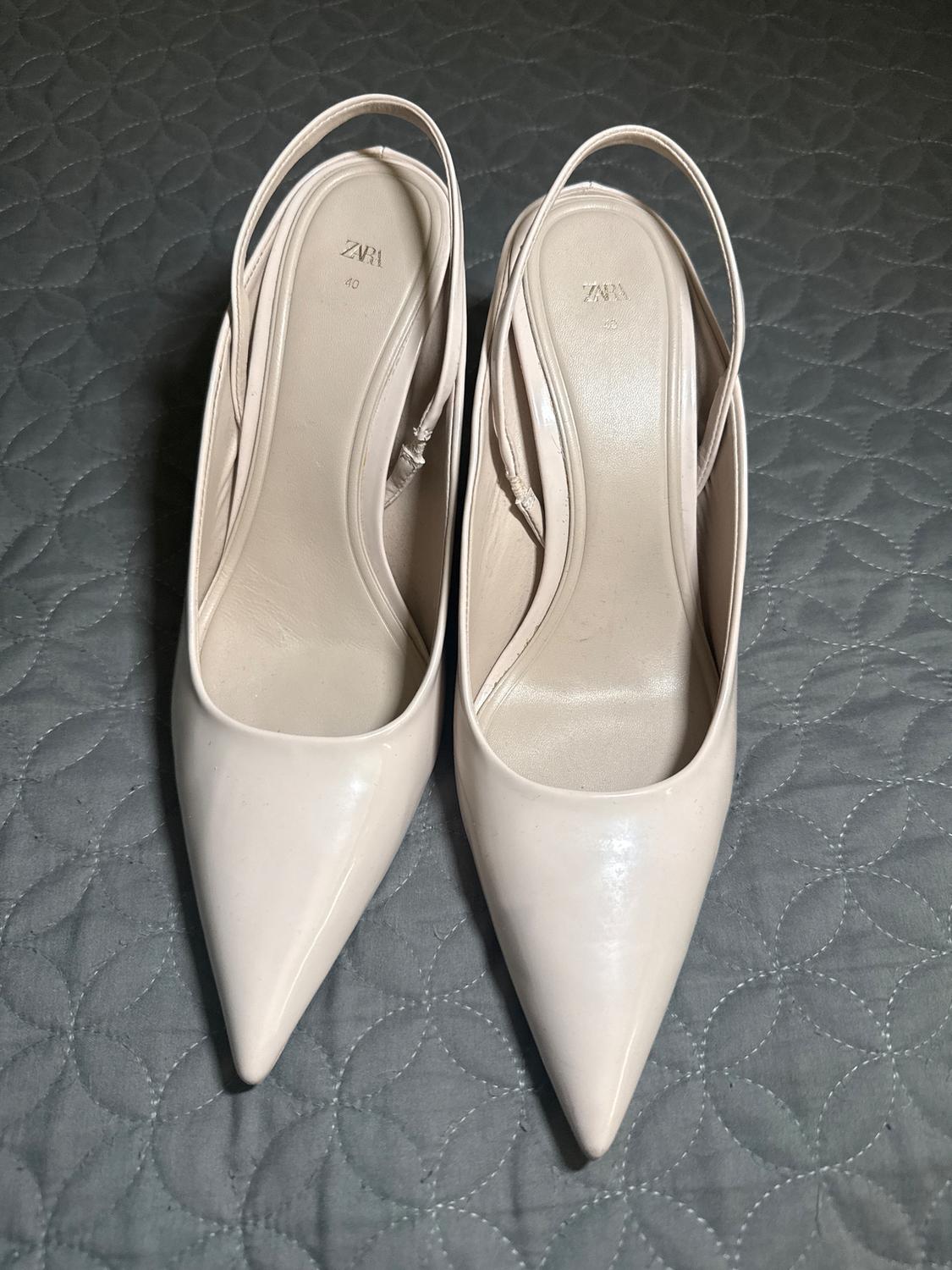 White pointed-toe slingback heels on a gray comforter with a circle pattern. Visible text: "ZARA 40".