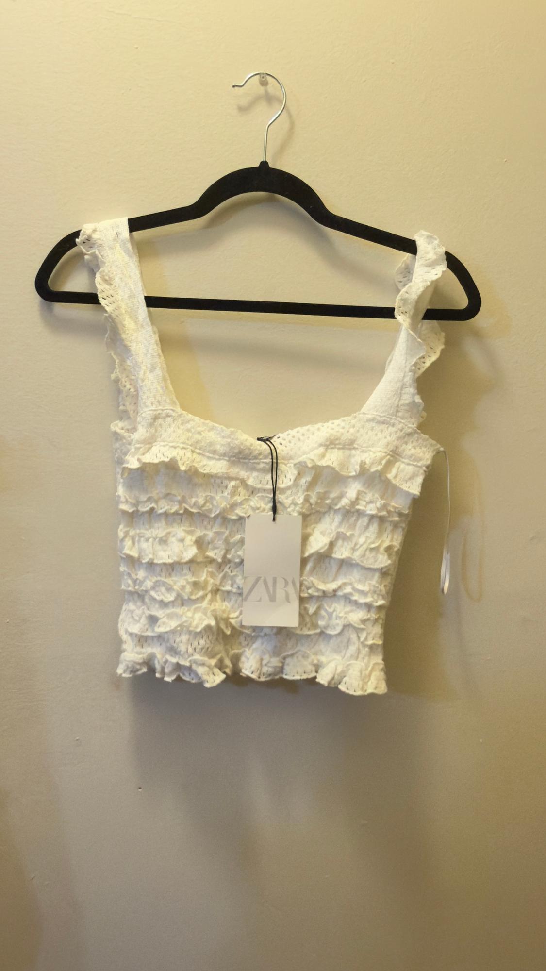 White ruffled top hanging on a black hanger. Visible label with text "ZARA". Beige background.