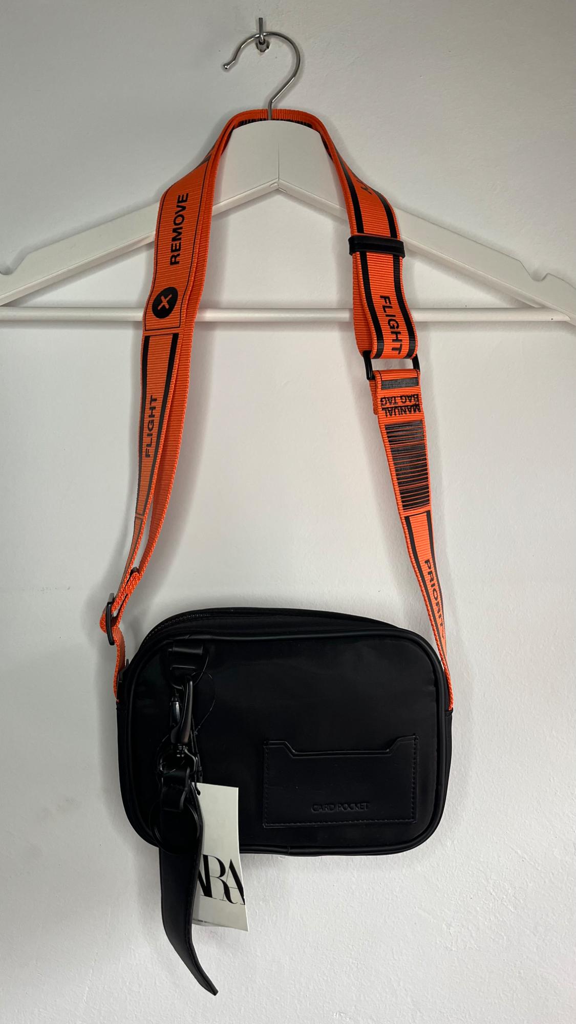 Black bag hanging on a white hanger with an orange strap featuring the text "REMOVE", "FLIGHT", "PRIORITY". White tag with black lettering.