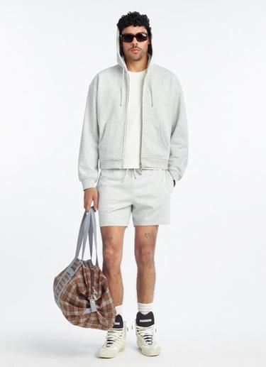 Modelo con sudadera gris con capucha, shorts a juego, camiseta blanca, gafas de sol, zapatillas blancas y bolsa de cuadros marrones.