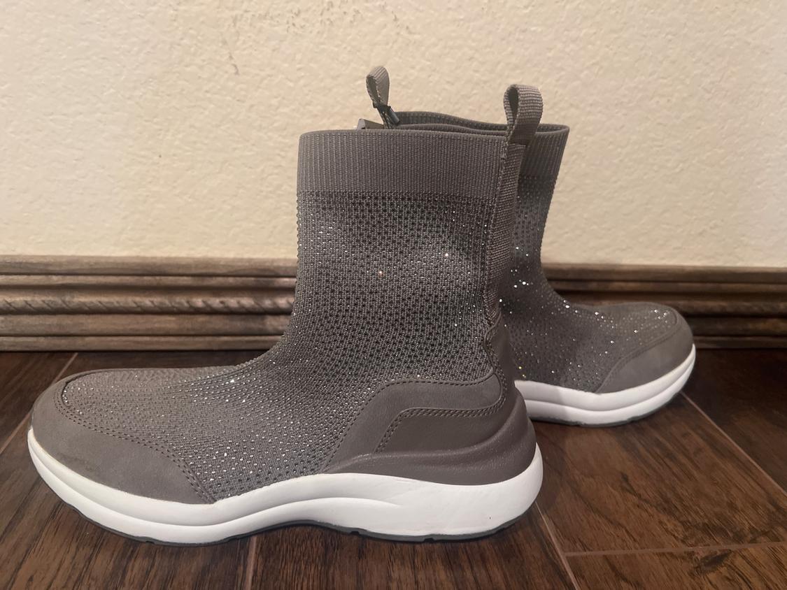 Grey boots with a shiny texture and white sole on a wooden floor. Beige wall background with wooden trim.