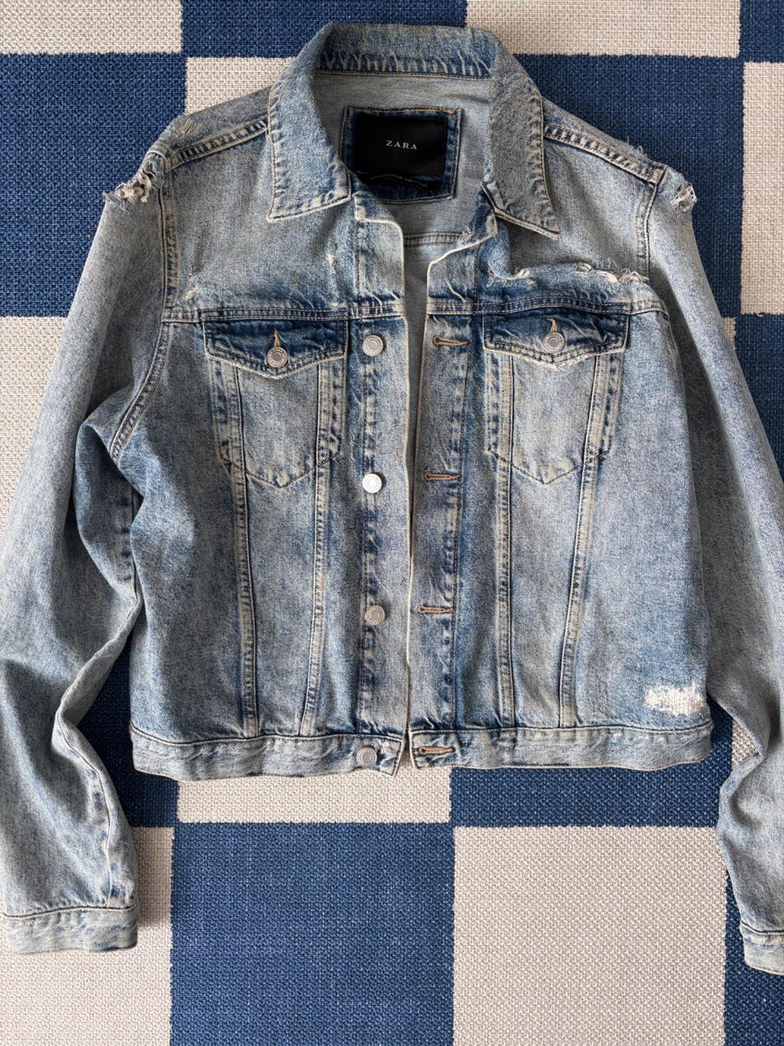 Distressed denim jacket with metal buttons over a blue and beige plaid background. Visible tag with text: ZARA.